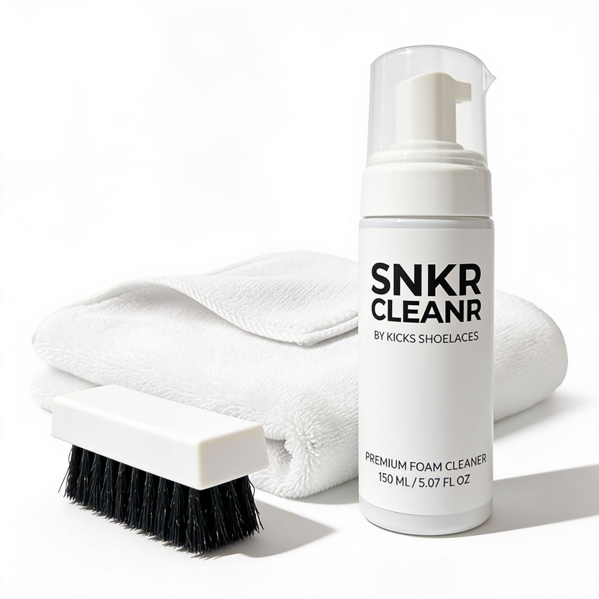 A white foam bottle of SNKR CLEANR premium foam cleaner stands in front of a folded white towel, with a small white brush that has black bristles placed to the left, all on a clean white background.