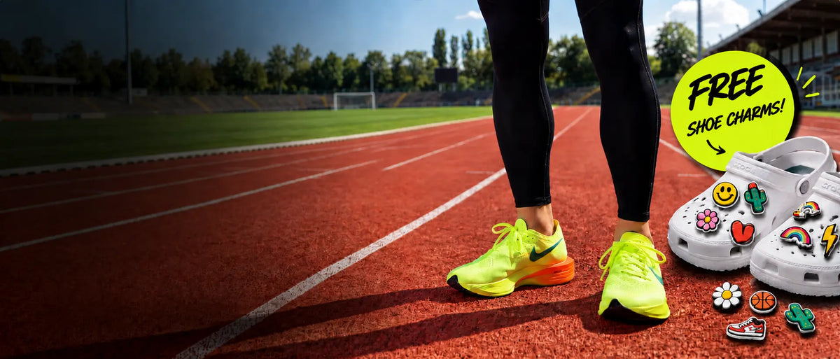 A person standing on a running track wearing bright neon yellow running shoes, with a pair of white clogs covered in colorful shoe charms displayed beside them. A bright yellow circular badge reads 'FREE SHOE CHARMS!'.